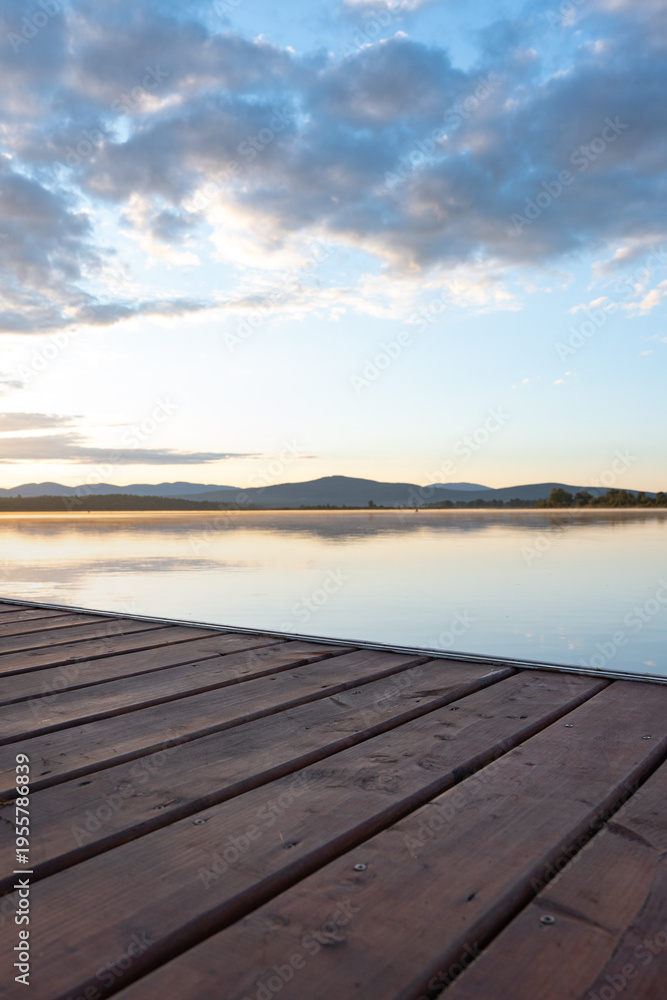 Fototapeta premium Vertical landscape of a serene lake at sunrise, viewed from a wooden deck in the foreground. Calm water reflects soft morning light, with mountains rising in the middle distance and colorful sky above