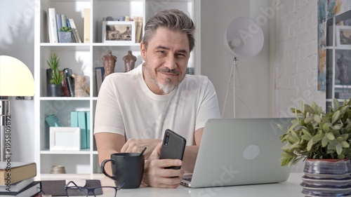 Older white man in glasses sitting at desk using phone. Home office, businessman working from home, morning coffee, checking social media news feed.