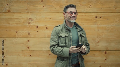 Man holding mobile phone - telecommunication and social media. Middle aged male outdoor in front of wooden wall wearing casual clothing. Eyeglasses and gray hair. Happy satisfied smiling.