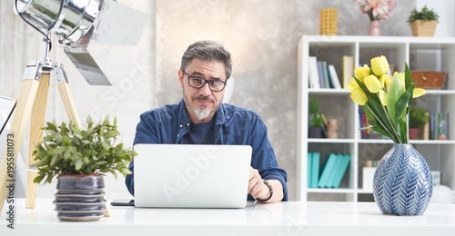 Bearded man working online with laptop computer at home sitting at desk. Home office, browsing internet, study room. Portrait of mature age, middle age, mid adult man in 50s.