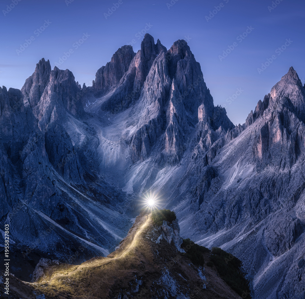 Naklejka premium Flashlight trails on mountain path against high rocks at night in autumn. Tre cime, Dolomites, Italy. Colorful landscape with light trails, trail on the hill, mountain peaks, sky with stars in fall