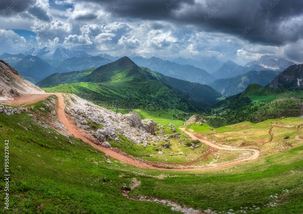 Obraz premium Winding mountain road in a vibrant green valley, alpine mountain peaks under a dramatic sky filled with contrasting light and shadow in summer. Panoramic colorful landscape. Dolomites, Italy. Nature
