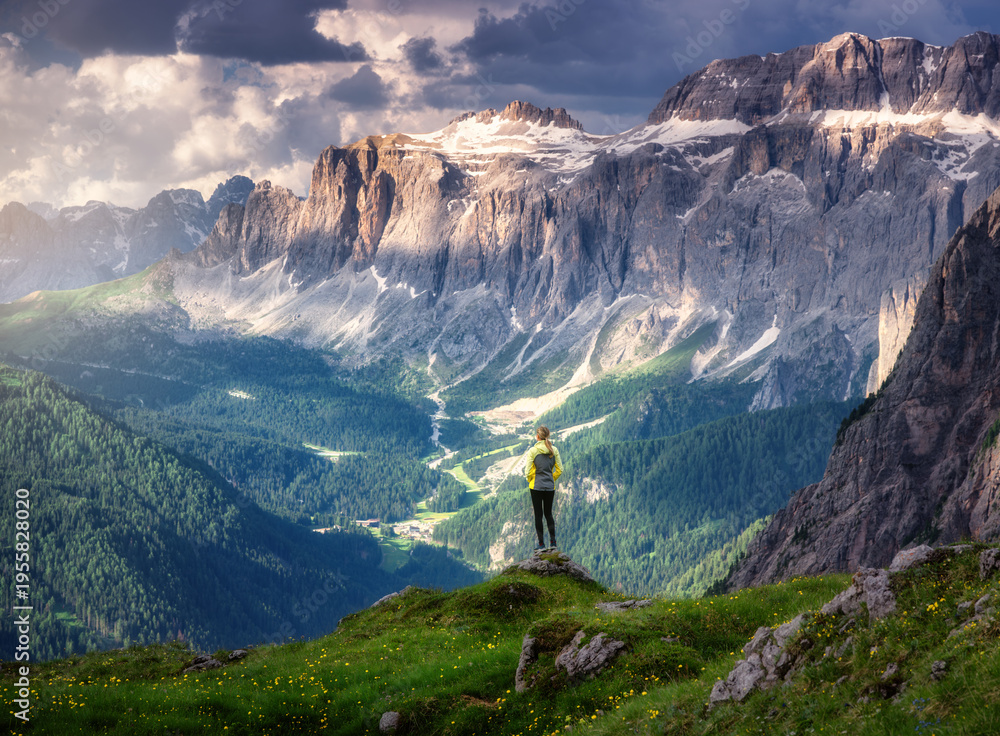 Fototapeta premium Sporty woman standing on a grassy hilltop with stunning view of the alpine mountains at sunset in summer. Scenery with girl, dramatic clouds over rocks and green valleys. Hiking in Dololmites, Italy
