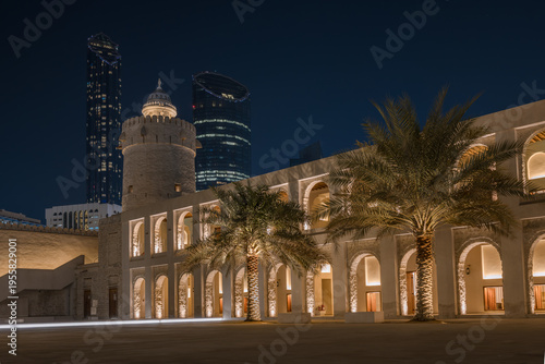 The historic Qasr Al Hosn fort illuminated at night with palm trees and arched arcade, contrasting with modern glass skyscrapers in the background, Abu Dhabi, United Arab Emirates 