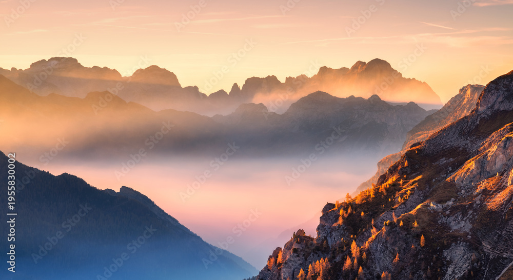 Fototapeta premium Mountains in low clouds at beautiful sunset in autumn in Dolomites, Italy. Landscape with alpine mountain hills in fog, orange trees and grass in fall, colorful sky with golden sunbeams. Aerial view