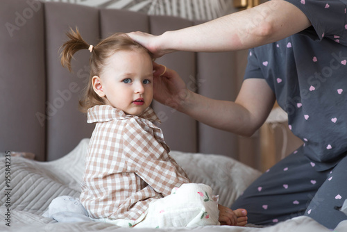 A happy mother is making pigtails on her little 2-year-old daughter's head while sitting in bed in the morning.