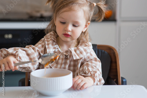 A 2-year-old girl in plaid pajamas is sitting at a table eating porridge from a white plate. Baby food. Feeding a baby. Healthy eating.
