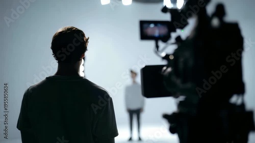A person is filmed in a minimalist studio with dramatic lighting, creating a moody and artistic scene. The camera setup and blurred background emphasize the filmmaking environment