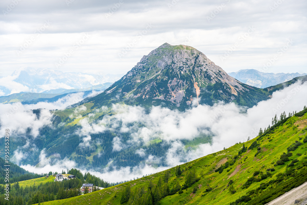 Fototapeta premium Majestic mountains rise above the rolling clouds in the Low Tauern region of Austria. Lush green hills contrast with rocky peaks, offering a stunning view of nature's beauty.