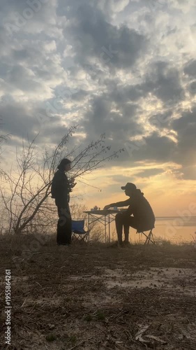 Silhouette of a couple having a picnic at sunset, sitting at a table outdoors, cloudy sky background. Man and woman drinking tea during an outdoor picnic, backlit silhouettes 