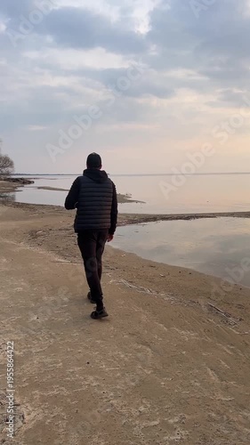 Rear view of a man walking on a deserted beach under a dramatic cloudy sky, peaceful nature scene. River side in early spring or winter. Mindful walk and peace moments