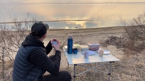 Outdoor picnic by the lake in cold weather, man sitting near camping table with hot drink and snacks. Calm water and nature background
