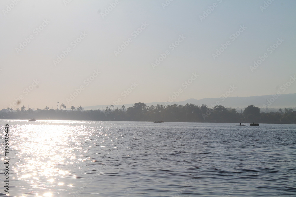 Naklejka premium Beautiful morning landscape of a wide tropical river with sunlight reflecting on the water surface and silhouettes of traditional boats near a lush palm tree coastline under a clear misty sky.