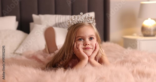 Beautiful little girl with crown dreaming about becoming queen on bed at home