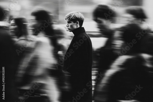 Young man standing still in crowded street with blurred people walking past, black and white portrait capturing urban life and isolation in motion.