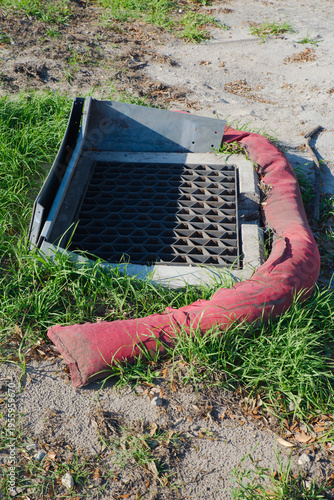Εκτύπωση καμβά A metal storm drain grate surrounded by a red silt sock on sandy soil and grass, illustrating erosion control, stormwater protection and roadside drainage maintenance at a construction or urban site