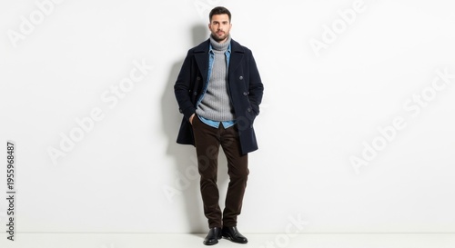 A man wearing a coat stands against a white wall with his hands in pockets.