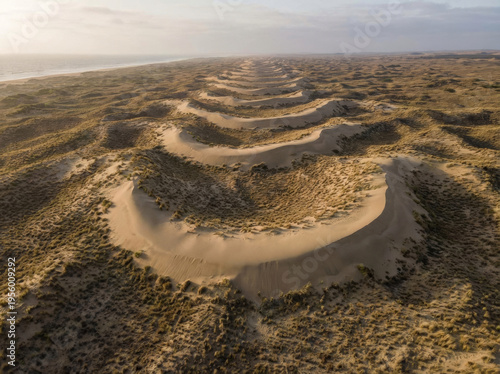 Expansive aerial view of rolling sand dunes covered in sparse vegetation leading towards the ocean horizon.