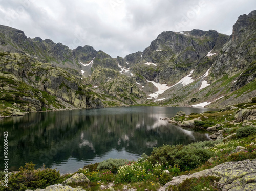 Tranquil mountain lake reflecting rugged peaks under a cloudy sky, surrounded by rocky terrain and green vegetation.