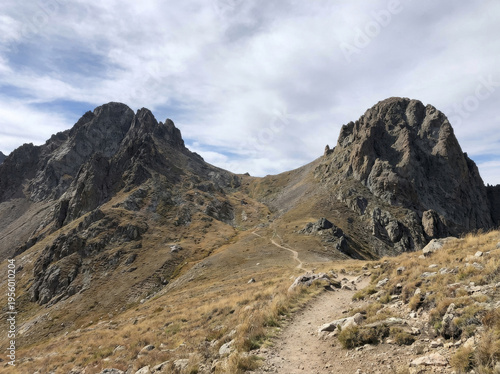 A winding hiking trail leads through dry grass towards rugged, rocky mountain peaks under a cloudy sky.
