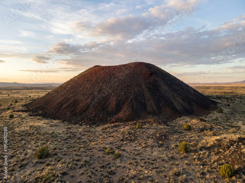 A solitary cinder cone volcano stands against a vast desert landscape under a dramatic sky.