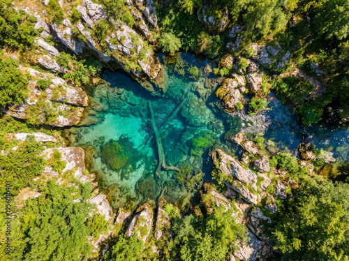 Aerial drone view of a crystal clear turquoise spring surrounded by lush green forest and rocky terrain.