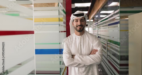 Confident young Middle Eastern business professional man in traditional Arabic dress standing in office corridor, keeping hands folded, looking at camera, getting happy, posing for head shot portrait