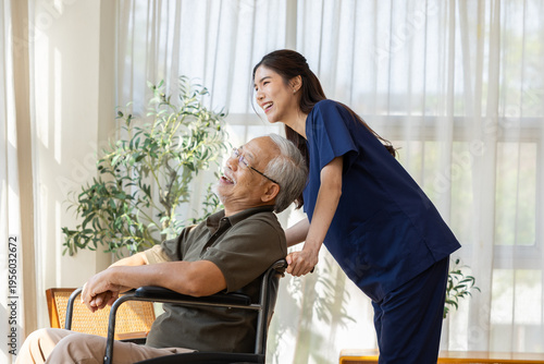 Happy elderly Asian man sitting in wheelchair smiling and laughing with young cheerful nurse caregiver near bright window indoors enjoying warm sunlight and emotional connection