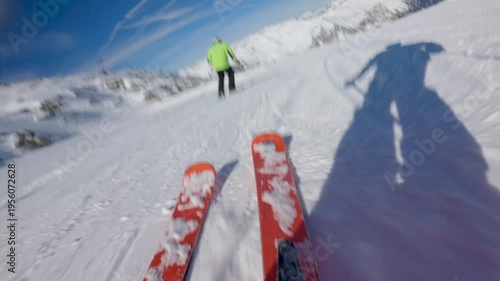 Skier in a green jacket skis down a snowy mountain. View from the perspective of the skier, showing their skis and shadow. Low angle slow motion
