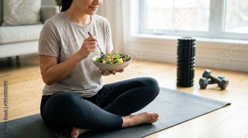Happy young woman eating healthy green vegetable salad smiling sitting on yoga mat in living room after fitness workout session enjoying natural organic diet and peaceful active lifestyle
