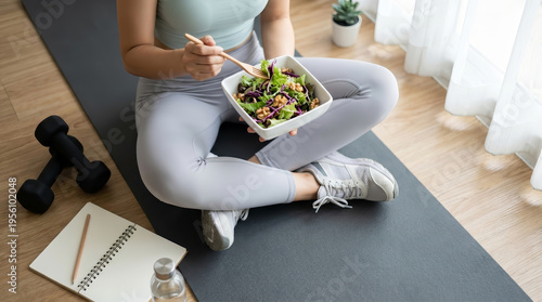 Active woman in sportswear sitting on yoga mat eating healthy green salad feeling relaxed after indoor home workout near dumbbell notebook water bottle