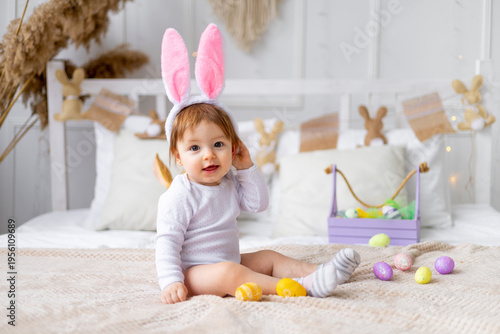 cute happy baby girl in bunny ears on the bed at home with a rabbit and painted eggs, easter concept, funny Easter baby is waiting for the holiday and rejoicing