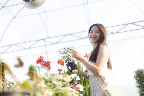Happy young asian woman smiling holding potted plant inside bright greenhouse nursery enjoying casual gardening hobby with cheerful joyful emotion surrounded by beautiful fresh green flora