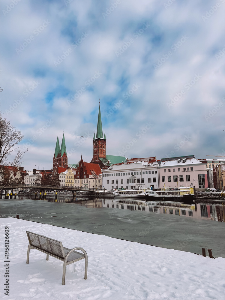 Obraz premium Frozen Trave river with snowy bench and Luebeck Old Town skyline in distance