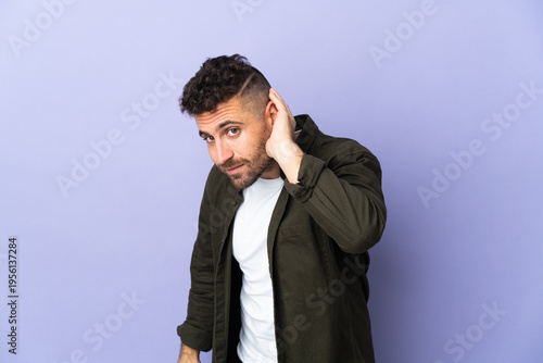 Caucasian man isolated on purple background listening to something by putting hand on the ear