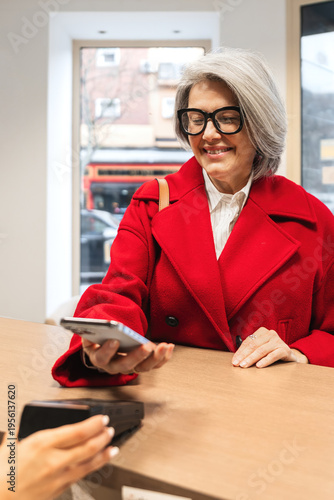 Mature woman in a red coat making a contactless payment using her smartphone at a reception counter in a modern clinic