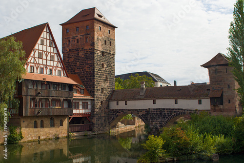 Nurnberg, Germany. Medieval towers and timbered house rise above the Pegnitz. Stone arches carry a covered bridge between towers. Green banks reflect warm roofs in calm water.