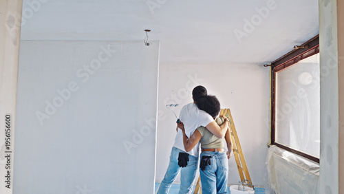 In a sunlit room under renovation, a couple paints the walls, sharing a moment of connection as they refresh their home's interior.