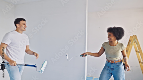 Fotografi In a sunlit room, a cheerful couple engages in playful banter while working on a home improvement project, with the man holding a paint roller and the woman wielding a paintbrush