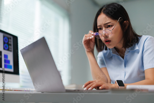 Foto Young Asian woman working in an office holding her glasses and looking at a lapt