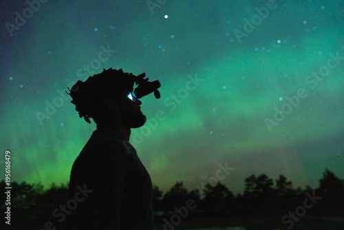 Profile silhouette of a man using night vision goggles to observe green Aurora Borealis under a starry northern sky.