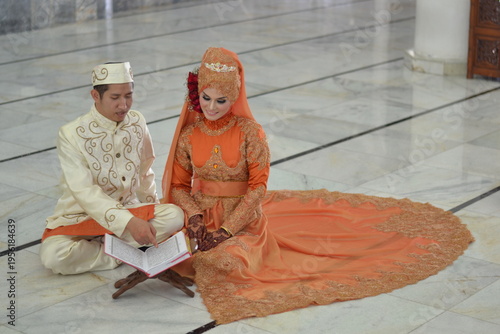 A Muslim couple in elegant traditional attire reading the Quran together on a marble floor inside a mosque after their wedding ceremony.
