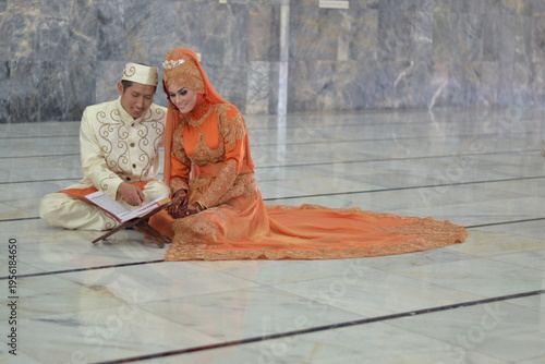 A Muslim couple in elegant traditional attire reading the Quran together on a marble floor inside a mosque after their wedding ceremony.