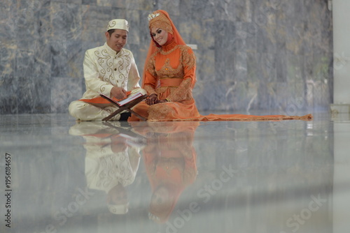 A Muslim couple in elegant traditional attire reading the Quran together on a marble floor inside a mosque after their wedding ceremony.