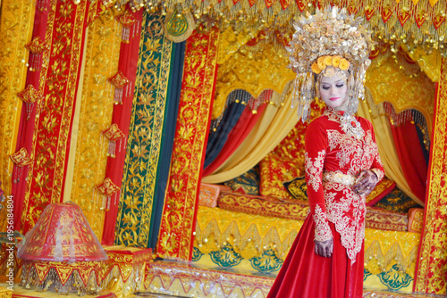 A portrait of a Minangkabau bride in a detailed red and gold traditional dress and elaborate headdress, posed against an ornate wedding backdrop