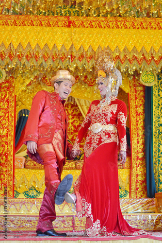 A stunning Acehnese couple in traditional red and gold wedding attire, posing elegantly against a vibrant, ornate ceremonial backdrop