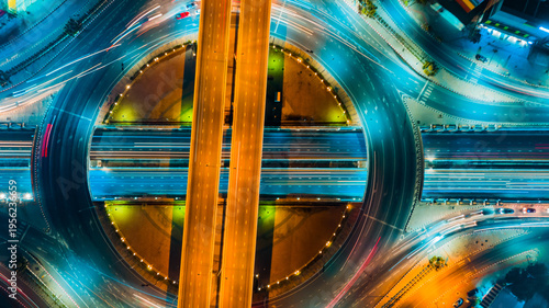 Aerial top view of diagonal highway crossing circular interchange at night, bright orange light trails slicing through glowing blue roads, captured with high angle and neon lighting,