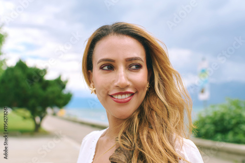 Close portrait of smiling African woman in her thirties outdoors by the sea, looking away with soft expression, natural beauty, long hair, white dress, fresh air and calm lifestyle mood