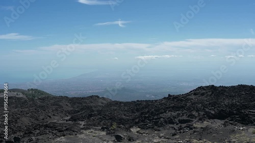 The camera pans right across black lava on Mount Etna with a hazy valley below and ends on a house beneath a blue sky and soft clouds.