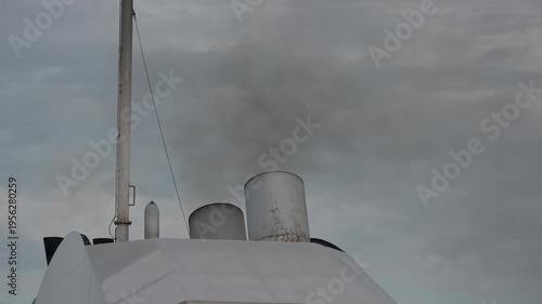 Close view of a large ship funnel with two exhaust openings releasing dense black smoke beside a vertical pole under dark cloudy sky.